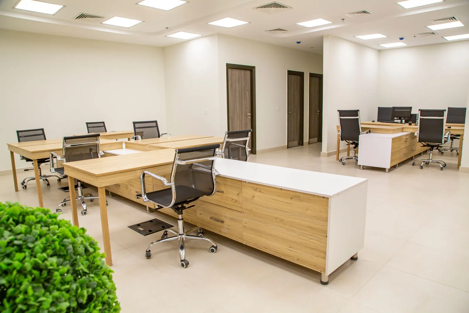 An empty open-plan office with light wood desks and black mesh chairs under bright ceiling lights