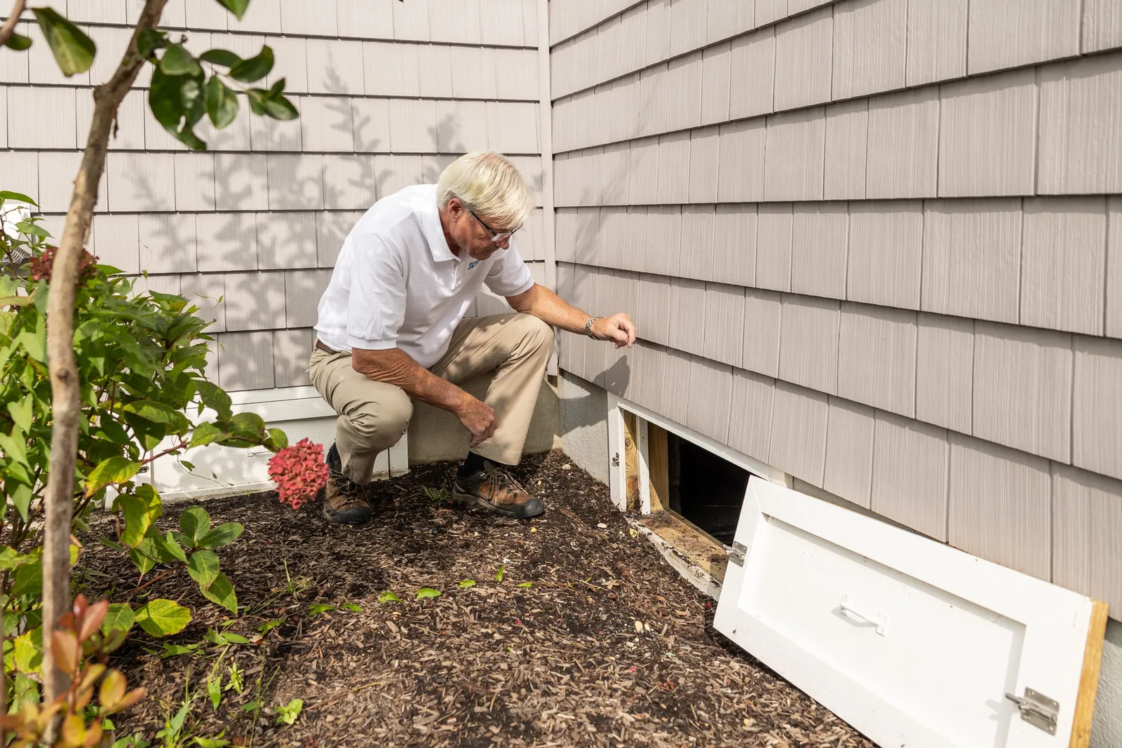 A man kneeling beside an open crawl space door on the side of a house