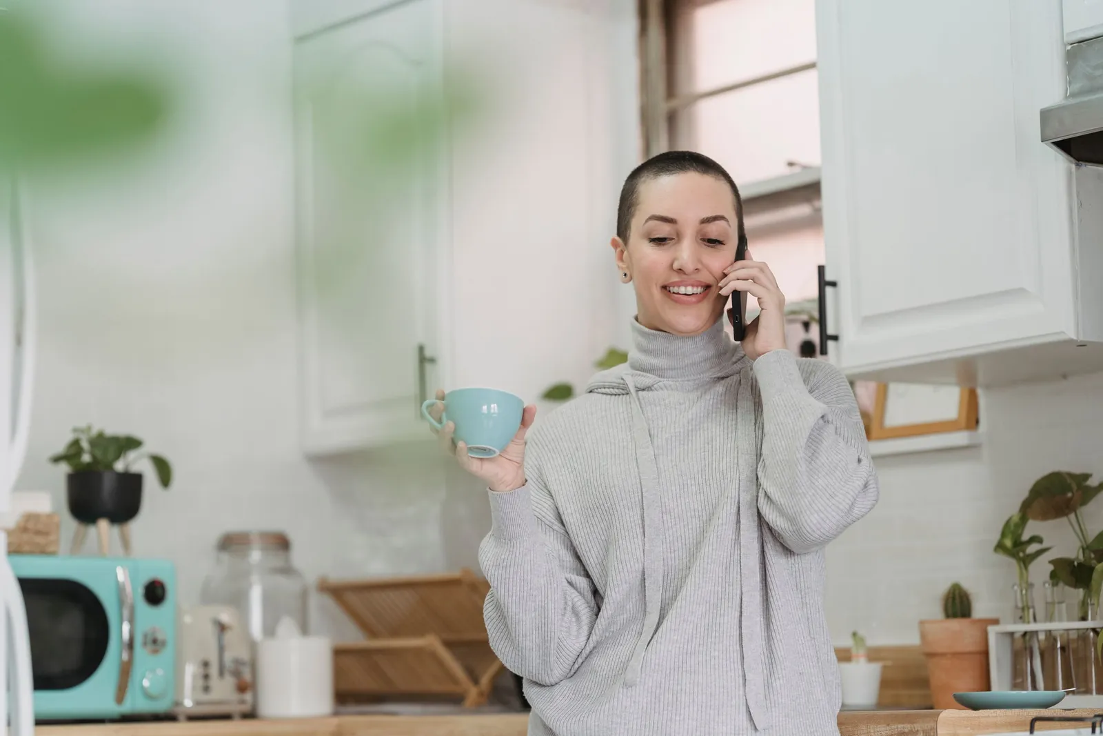 A woman in a gray sweater talking on the phone in her kitchen, holding a coffee cup