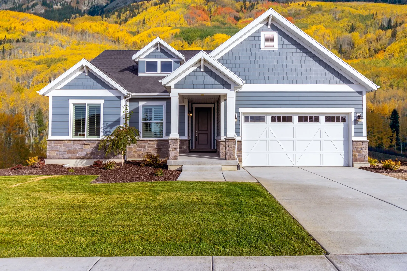 A blue craftsman-style house with a white garage door and yellow autumn trees on the hillside behind it